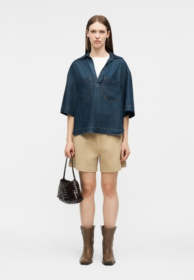 Femme portant une chemise en jean oversize bleu foncé, un short beige, des bottines marron, tenant un petit sac à main tressé foncé, debout sur un fond uni.