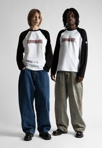 Due persone che indossano camicie a maniche lunghe bianche e nere coordinate con la scritta "Wasted", abbinate a jeans larghi e scarpe nere, in piedi affiancate.