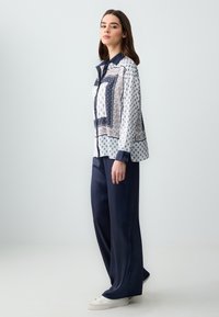 Blouse en soie avec un design à motifs en marine et blanc ; présente un devant boutonné, un col et des poignets contrastés. Associée à un pantalon large marine.