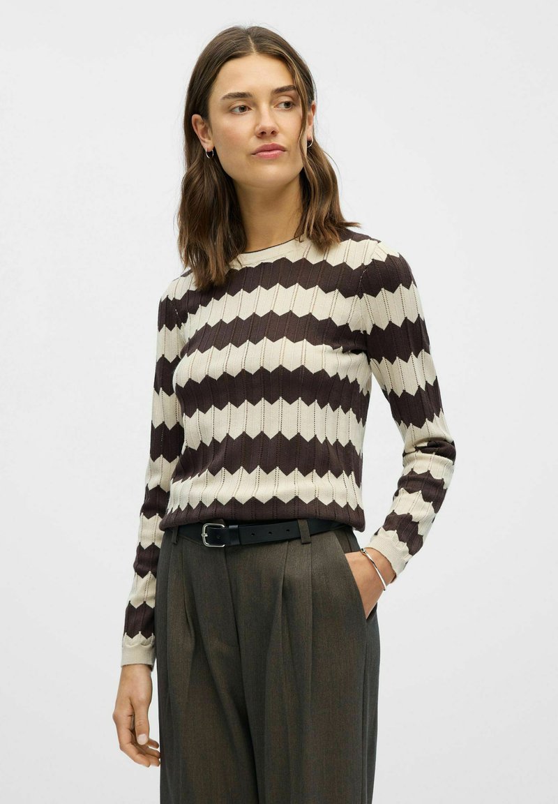 Cremfarbener und brauner Zickzack-Musterpullover mit langen Ärmeln und rundem Ausschnitt, kombiniert mit grauen, plissierten Hosen und einem schwarzen Gürtel.