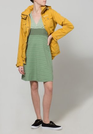 Mujer vestida con un vestido verde estampado debajo de una chaqueta amarilla desabrochada, con zapatillas negras, de pie con una mano en la cadera sobre un fondo liso.