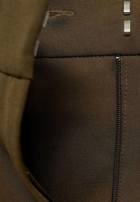 Tissu marron foncé avec une texture subtile ; comporte une fermeture éclair sur le côté droit et des accents métalliques près du haut.