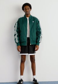 Groene bomberjack met witte strepen op de mouwen, gedragen over een wit T-shirt en zwarte shorts. Zwarte sneakers met witte accenten maken de look af.