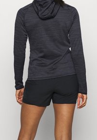 Felpa nera a maniche lunghe con tessuto testurizzato e design aderente, abbinata a pantaloni corti sportivi neri. Presenta un cappuccio e aperture per il pollice.