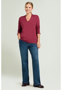 Bordeauxrode v-hals trui van zacht materiaal met lange mouwen, gecombineerd met blauwe wijde jeans en glanzende donkere schoenen. Subtiele zijsplitten aan de zoom.