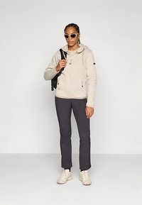 Felpa con cappuccio beige con zip e coulisse, tessuto texturizzato; pantaloni grigio scuro; sneakers bianche; zaino nero; occhiali da sole; posa in piedi.