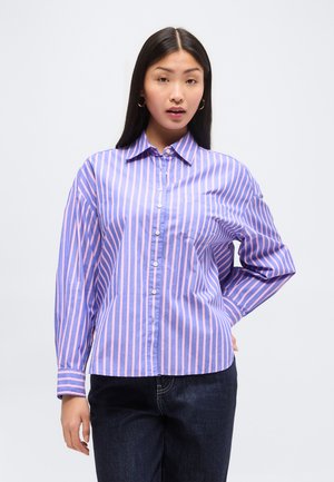 Mladá žena oblečená v modro-ružovej vertikálne pruhovanej košeli na gombíky a tmavých džínsach, stojaca s jednou rukou na bokoch.