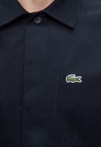Chemise bleu marine à col avec un bouton noir et un petit logo vert de crocodile brodé sur la poitrine gauche.