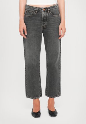 Forfra bøjle af person iført sorte falmede jeans med lige ben og sorte ballerinaer, stående mod en ensfarvet hvid baggrund.