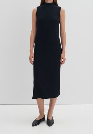 Woman standing wearing a sleeveless, ankle-length black dress with a high neckline and black pointed flats.