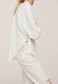 Weiße oversized Bluse mit langen Ärmeln, seitlichen Bindetails und lockerer Passform. Kombiniert mit hellen, ausgewaschenen Jeans. Zarte Textur.