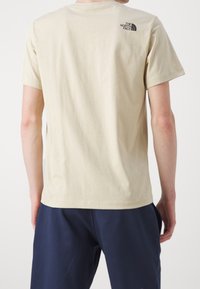 Camiseta de algodón beige con mangas cortas, cuello redondo y un pequeño logo negro en la parte superior de la espalda. Diseño simple y casual.