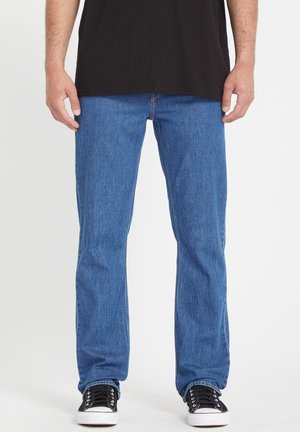 Homme portant un jean bleu à jambes droites, des baskets noires à semelles blanches, et un t-shirt noir, debout devant un fond uni.