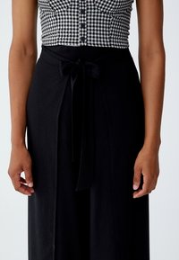 Zwarte wijde broek met een strikdetail in de taille, gemaakt van een gladde stof. De bovenkant heeft een zwart-wit ruitpatroon met knopen.