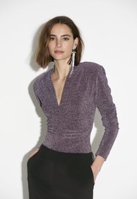Lila glitzerndes Oberteil mit langen Ärmeln, V-Ausschnitt und gerafften Seiten, kombiniert mit schwarzen Trousers. Bemerkenswerter Glanz und elegantes Design.