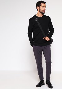 Črn dolg rokav pulover, slim-fit sivi hlače, usnjeni čevlji in torbica čez ramo. Modell stoji z eno roko v žepu, nevtralna ozadje.