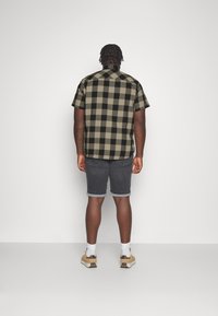 Camisa de cuadros en negro y verde oliva de manga corta, combinada con pantalones cortos grises con dobladillo enrollado y zapatillas color marrón claro, vista desde atrás.