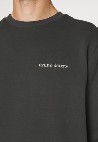 Felpa grigia scura con scollatura tonda. Presenta un piccolo logo bianco "LYLE & SCOTT" ricamato sul petto. Tessuto liscio.