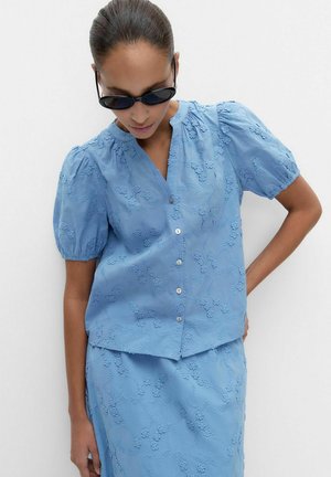 Femme portant un chemisier brodé bleu clair assorti avec manches bouffantes et jupe, accompagné de lunettes de soleil ovales noires, sur fond blanc.