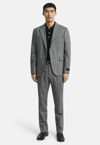 Grau angezogener Wollanzug mit Spitzrevers, einreihiger Blazer, glatte Vorderhose, getragen über einem schwarzen Poloshirt, kombiniert mit schwarzen Schuhen.