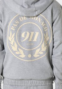 Sweat à capuche gris clair avec texte beige "Pas de Monaco 911 Club" et motif de couronne imprimés au dos.