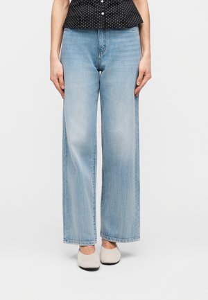 Kvinne iført lyseblå vide jeans, kremfargede flate sko og en svart topp med hvite prikker, står mot en ensfarget bakgrunn.