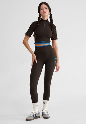 Crop top marron à côtes avec des manches courtes et un col haut, associé à des leggings taille haute assortis avec des accents bleus.
