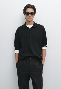 Schwarzes Strick-Polohemd mit drei Knöpfen, über einem weißen Hemd getragen. Dunkle Hose, Hände in den Taschen. Accessoires sind dunkle Sonnenbrillen.