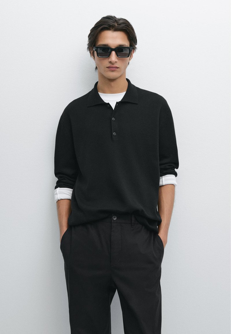 Polo shirt nero a maglia con tre bottoni, indossato sopra una camicia bianca. Pantaloni scuri, mani nelle tasche. Gli accessori includono occhiali da sole scuri.