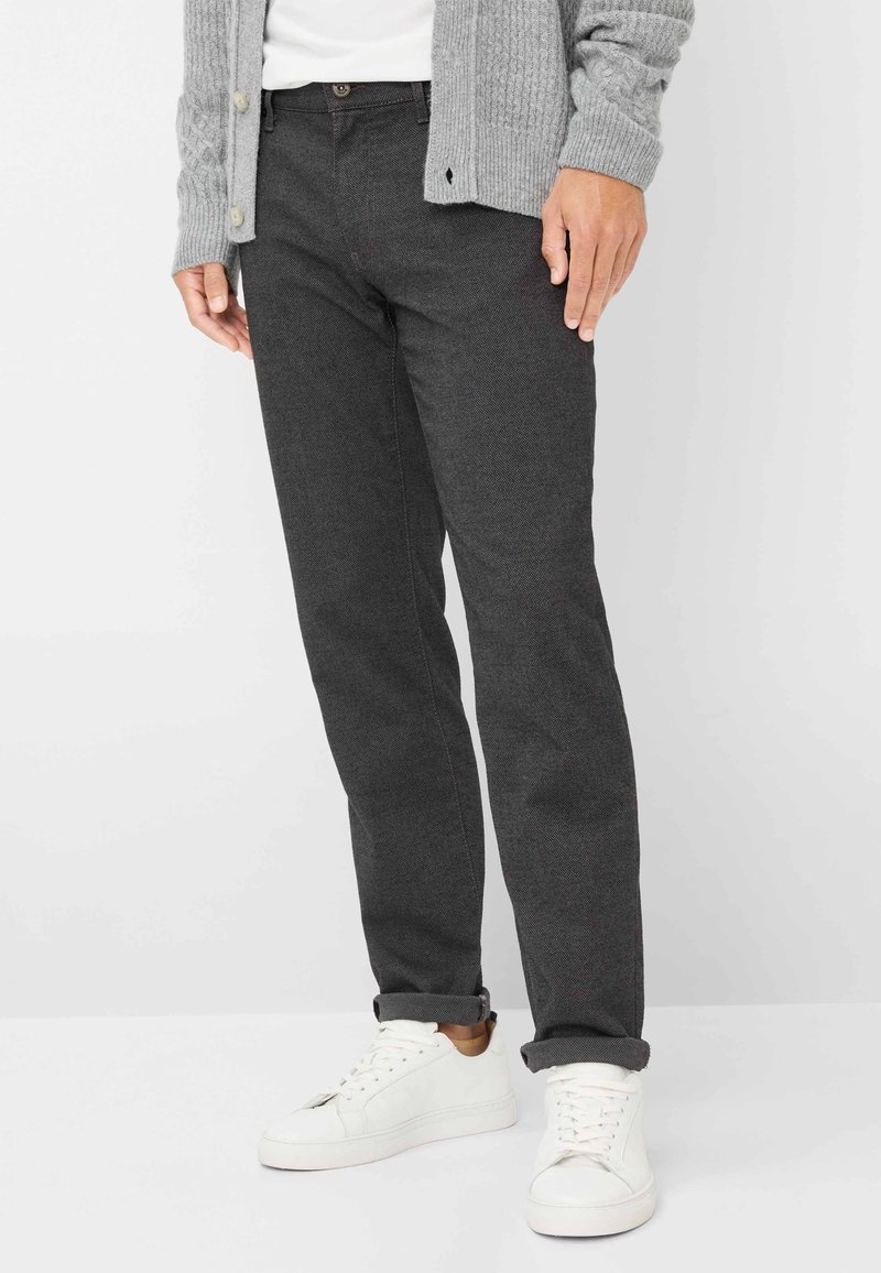 Homme portant un pantalon gris foncé, des baskets blanches, une chemise blanche et un cardigan en tricot gris, les mains détendues le long du corps.