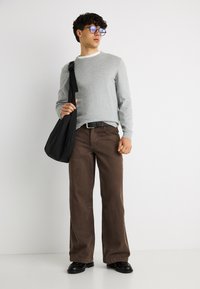 Jeune homme portant des lunettes, un pull gris, un pantalon large marron, une ceinture noire, des chaussures noires, tenant un sac à bandoulière noir, debout contre un mur blanc.