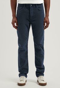 Pantalones de pana azul marino con un diseño de pierna recta. Presentan bolsillos frontales, cierre con botón y una textura sutil. Combinados con zapatillas blancas.