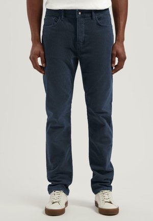 Pantalones de pana azul marino con un diseño de pierna recta. Presentan bolsillos frontales, cierre con botón y una textura sutil. Combinados con zapatillas blancas.