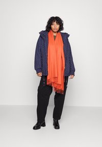 Navy polka dot jacket with hood, orange scarf with fringes, black pants, and black heeled ankle boots. Neutral background.