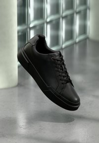 Μαύρο δερμάτινο αθλητικό παπούτσι χαμηλού προφίλ με κορδόνια, αιωρούμενο πάνω από γυαλιστερό γκρι πάτωμα, με υφή από γυάλινους τούβλους και λευκή κολώνα στο φόντο.