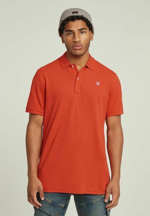 Polo di colore rosso con colletto e maniche corte. Presenta un piccolo logo sul petto. Il tessuto sembra liscio e leggero.