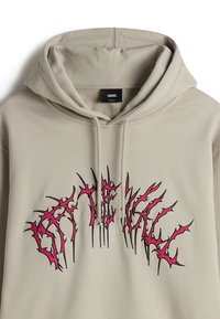 Beige hoodie met een grote, rode en zwarte grafische tekst "GATEWALL" in een doornenachtig, scherp lettertype. Heeft een capuchon met koordjes en een zachte textuur.