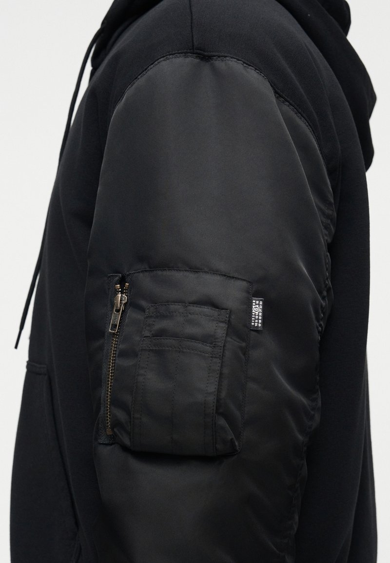 Sort hættetrøje med ærme i kontrastfyldt quiltet design, udstyret med lynlåslomme og mærkat. Glat stofoverflade med mat finish.