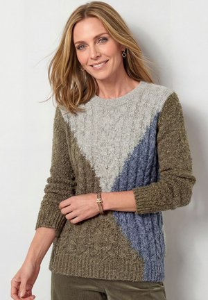 Glimlachende vrouw met blond haar draagt een gestructureerde gebreide trui in groen, grijs en blauw met een geometrisch patroon en een armband om haar pols.