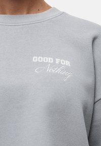 Felpa grigia chiara con colletto a costine. Presenta una scritta ricamata in bianco che recita "GOOD FOR Nothing" in stili di carattere misti.