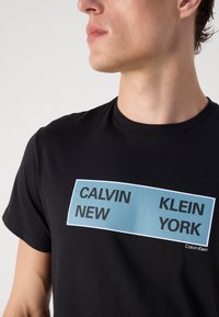 Juodas medvilninis marškinėliai su stačiakampiu šviesiai mėlynu grafiniu atvaizdu, kuriame didžiosiomis juodomis raidėmis parašyta "CALVIN NEW KLEIN YORK". Trumpomis rankovėmis ir apvaliu kaklu.