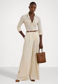 Bežinė marškinė su apykakle ir dviem krūtinės kišenėmis, derinama su grietinėlės spalvos plačiais kelnais. Rudas diržas ir rankinė, abi su tekstūrinėmis paviršiais.