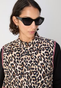 Μαύρα καμπυλωτά τρίγωνα γυαλιά ηλίου συνδυασμένα με ένα επενδυμένο γιλέκο με λεοπάρ σχέδιο που διαθέτει κουμπιά και ένα μαύρο μπλουζάκι με μακρύ μανίκι από κάτω.