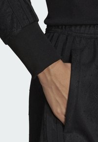 Svart träningskläder med ett texturerat, mönstrat tyg och ribbad mudd. Handen är placerad i en ficka, vilket visar en figurnära silhuett.