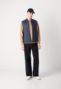 Colete acolchoado azul-marinho sobre um sweatshirt bege, combinado com jeans largos pretos e ténis brancos. O colete possui um fecho e um bolso.