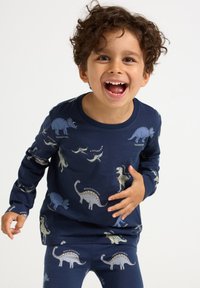 Smiling young boy with curly hair wearing navy blue pajamas patterned with various dinosaurs, standing against a plain white background.