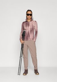 Majica dugih rukava s patentnim zatvaračem u različitim nijansama mauve, s rebrastom teksturom, uparen s svijetlo bež hlačama i smeškim planinarskim čizmama, drži trekarske štapove.