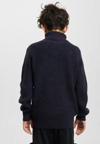 Tumši zili adīti džemperi ar augstu apkakli, teksturētu audumu, ribotām aprocēm un apakšmalu. Aizmugures skats parāda brīvu piegriezumu un vienkāršu dizainu.