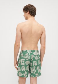 Πράσινα σορτς κολύμβησης με άσπρο αφηρημένο σχέδιο, ελαστική μέση και πλαϊνές τσέπες. Το ύφασμα φαίνεται απαλό με ελαφριά υφή.