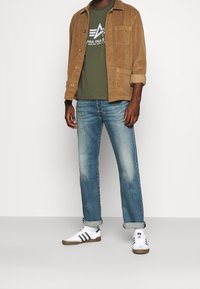 Hnědá kordová bunda, olivově zelené tričko s logem, modré vybledlé džíny a bílé tenisky se černými pruhy. Podrobnosti o neformálním outfitu.